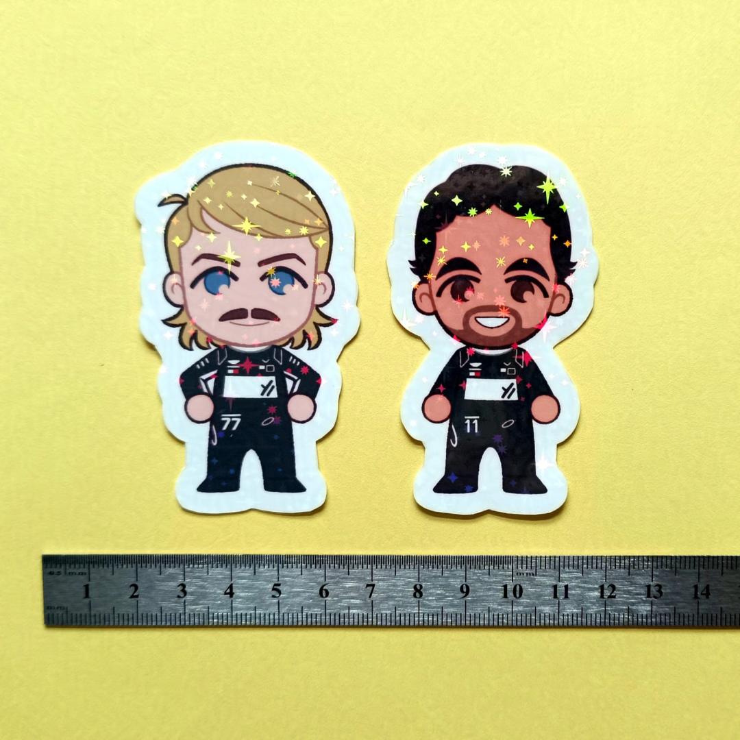 Two holographic stickers of chibi Valtteri Bottas and Sergio Checo Perez with a centimetre ruler for scale against a yellow background.