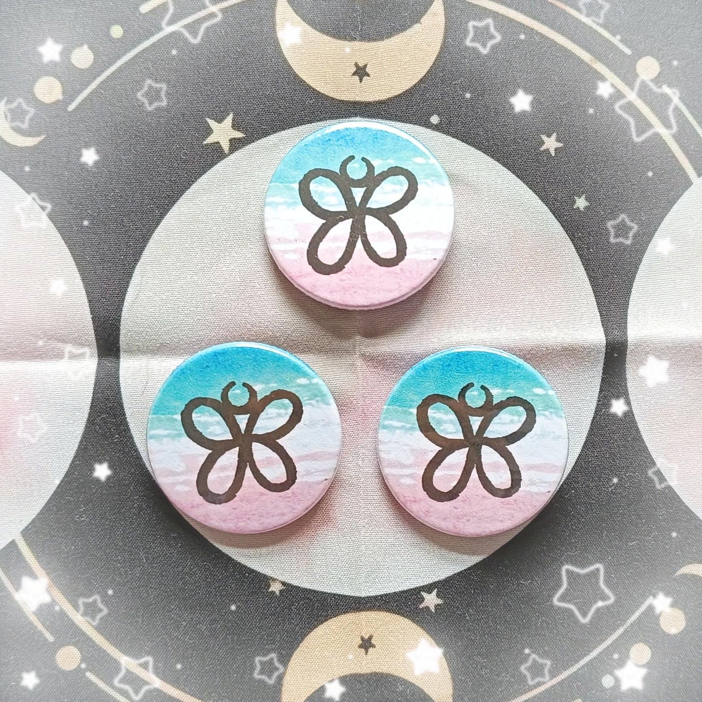 Photo of 3 Trans Rights Sigil Button Badges. The sigil looks like a butterfly. The sigil is in front of a blue, white and pink watercolour background.