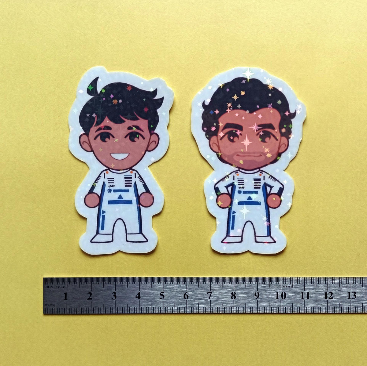 Two holographic stickers of chibi Alexander Albon and Carlos Sainz Jr, with a centimetre ruler for scale against a yellow background.