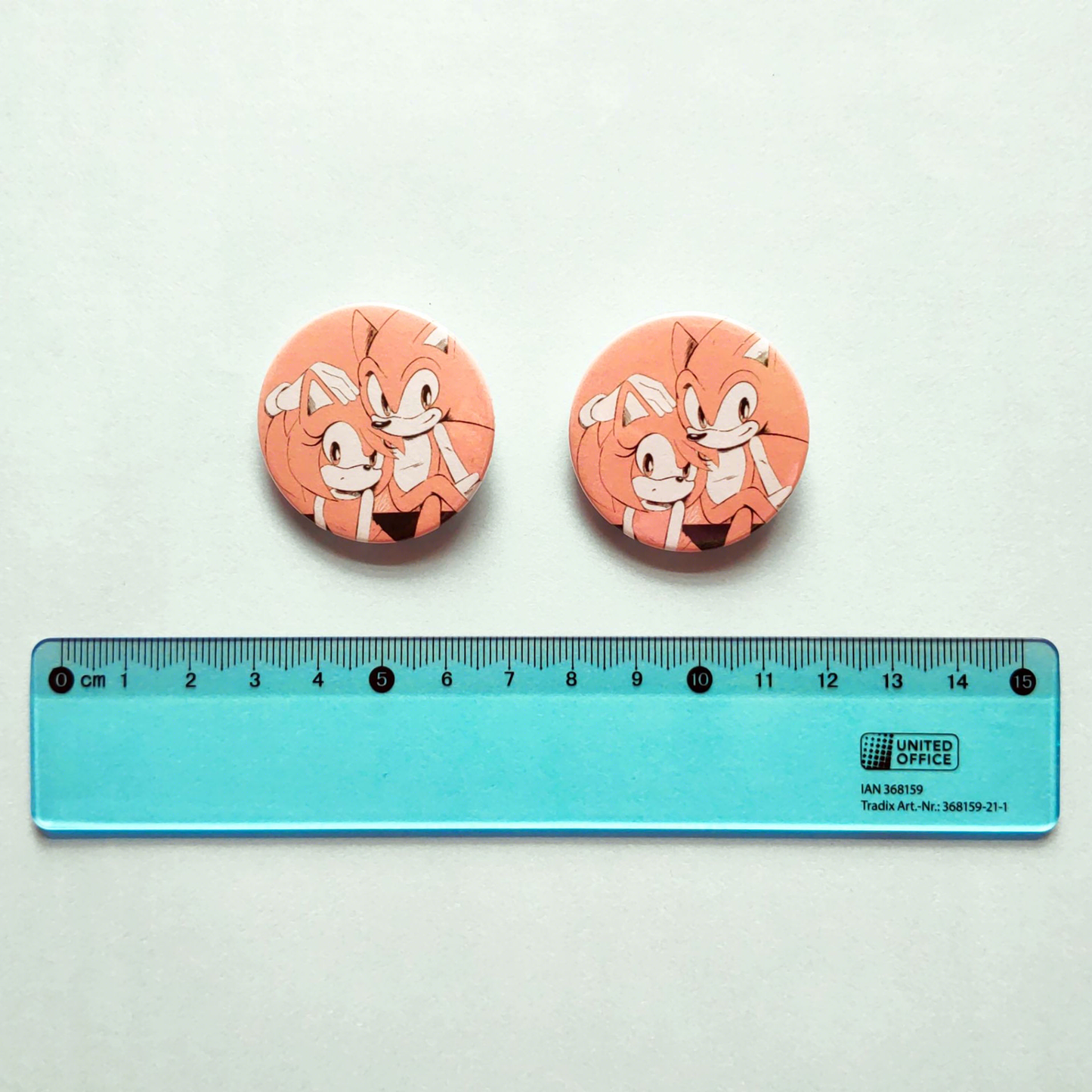 Photo of a pink Sonic and Amy button badges laid across a blue background, with a blue transparent ruler next to it to show size comparison.