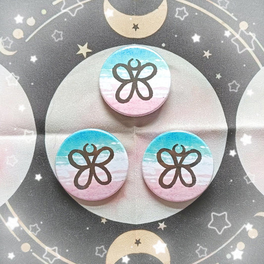 Photo of 3 Trans Rights Sigil Button Badges. The sigil looks like a butterfly. The sigil is in front of a blue, white and pink watercolour background.